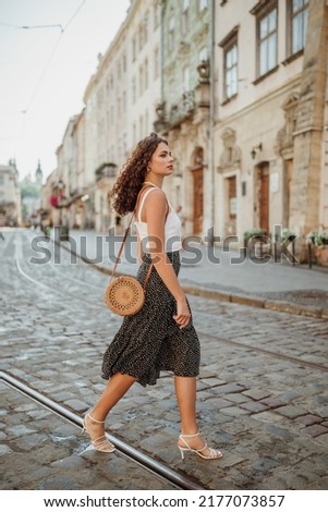 Elegant curly brunette woman wearing trendy summer outfit with round wicker shoulder bag, white top, polka dot midi skirt, strap sandals, walking in street of European city. Fashion, lifestyle concept Royalty-Free Stock Photo #2177073857