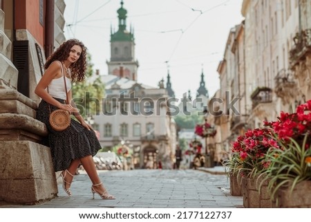 Elegant curly brunette woman wearing trendy summer outfit with round wicker shoulder bag, white top, polka dot midi skirt, strap sandals, posing in street of European city. Copy, empty space for text Royalty-Free Stock Photo #2177122737
