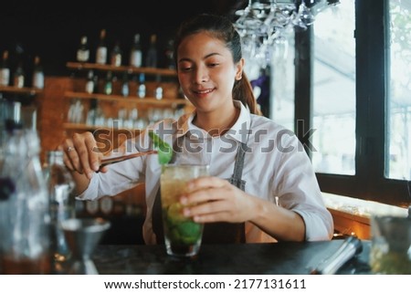 Beautiful Asian woman in gray apron preparing cocktail on the bar counter. Royalty-Free Stock Photo #2177131611