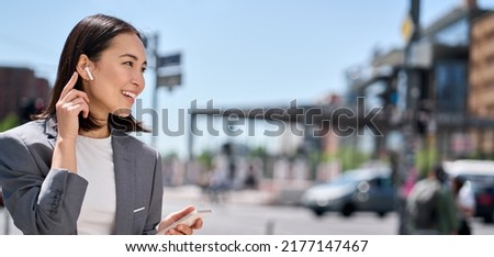 Young professional Asian business woman wearing suit using wireless earphones mobile internet data holding smartphone talking on mobile phone having chat on cellphone walking on city street. Banner Royalty-Free Stock Photo #2177147467
