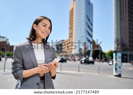 Young smiling professional Asian business woman entrepreneur wearing suit holding smartphone looking at mobile phone using apps on cellphone tech ordering taxi standing on urban city street. Royalty-Free Stock Photo #2177147489