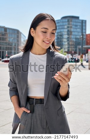 Young smiling elegant Asian busy business woman leader wearing suit standing in big city using cell phone platform applications. Smiling woman holding smartphone walking on street outdoors. Royalty-Free Stock Photo #2177147493