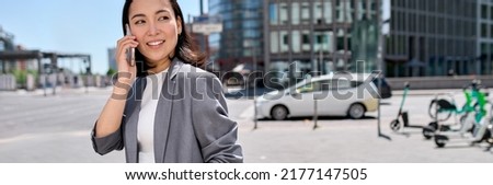 Young happy Asian successful businesswoman entrepreneur wearing suit standing in big city talking on mobile phone. Smiling woman making business call on cell walking on busy street outside.