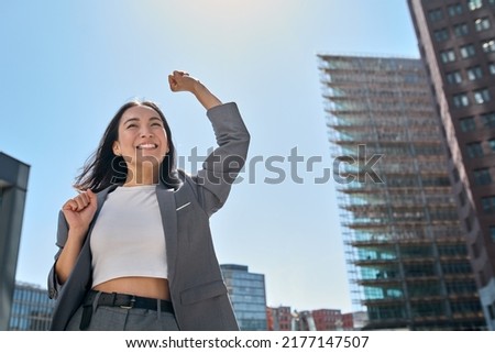 Young excited confident proud Asian business woman winner wearing suit standing on street, raising hands, feeling power, motivation, energy, celebrating career financial success in big city outdoors. Royalty-Free Stock Photo #2177147507