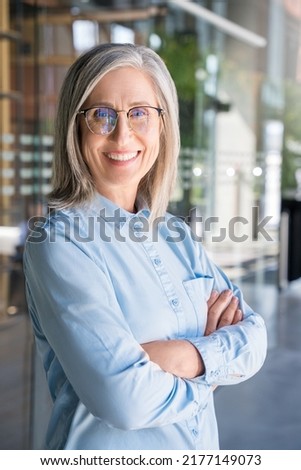 Vertical portrait of 60s gray-haired lady, old senior businesswoman, business leader manager looking at camera arms crossed. Smiling confident stylish mature middle aged woman standing in office.