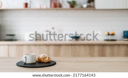 Croissant and coffee on kitchen countertop, against blurred minimalist interior with modern furniture. Selective focus at homemade pastry and tea drink in cup on wooden table, copy space, web banner Royalty-Free Stock Photo #2177164143