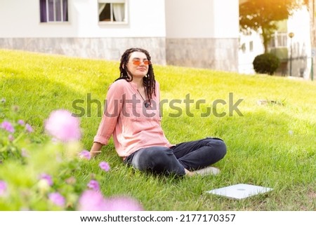 Photo of european woman 25s sitting on green grass in park with legs crossed during summer day while using laptop. Caucasian female hipster with dreadlocks and pink sunglasses on use laptop freelancer