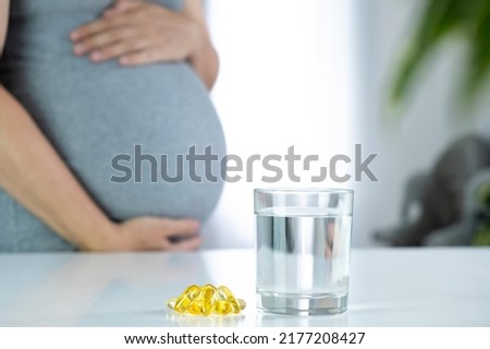 Omega 3 capsules and glass of water and pregnant woman at home interior. Healthy fatty acids nutritional supplement for prenatal support. Omega, DHA, vitamin D, fish oil for healthy pregnancy. Royalty-Free Stock Photo #2177208427