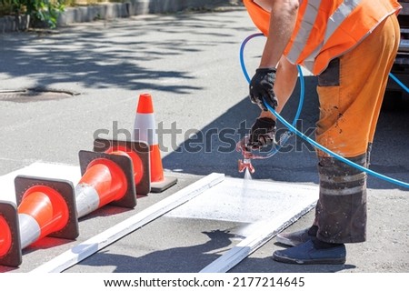 A road worker using a paint sprayer applies white road markings to a crosswalk using a wooden template. Copy space.