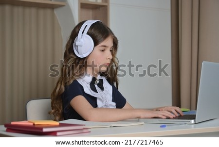 School girl in school uniform wearing wireless headset or headphones, sitting at table, studying from home in front of laptop and looking at computer screen.

Online learning and e-education concept.