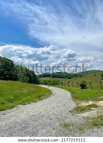 Female Hiker On Path Walking on Gravel Trail at The Moses Cone Memorial Park Off of Blue Ridge Parkway in Appalachian Mountains on Beautiful Sunny Day With Blue Sky and White Clouds Next to Farm