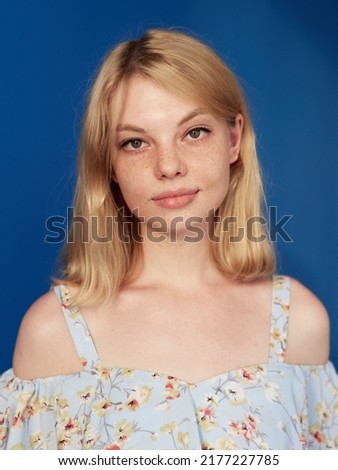 Closeup front image of a blonde young woman with blonde hair, looking at camera, isolated blue background.