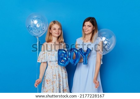 Two happy young women, posing in a studio, holding blue balloons and balloon with the inscription boy, blue background.