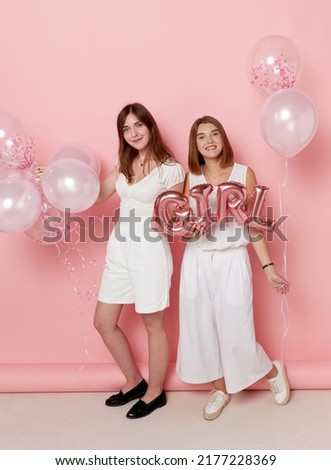 Full length image of a two of young friends, dressed in a white, holdings a balloons, over pink background.