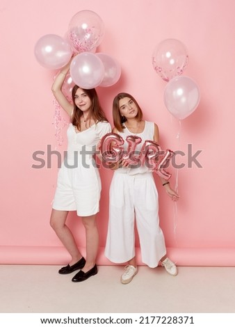 Full length view of smiling two girl, dressed in a white, holdings a balloons, over pink background.