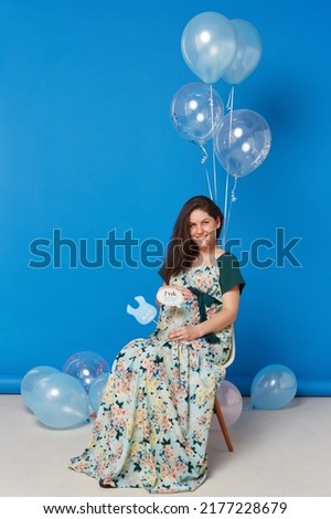 Full length image of a pregnant cheerful brunette young woman sitting on the chair with balloons, on blue background.