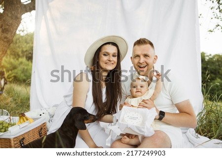 Positive married couple sitting with their dog and cute little daughter on white blanket with delicious cake. Happy family celebrating birthday outdoors with beautiful decoration around.