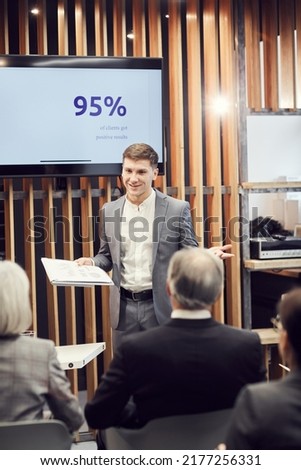 Smiling confident young businessman in suit standing in front of audience and presenting successful marketing cases at business conference Royalty-Free Stock Photo #2177256331