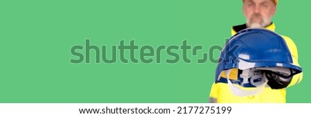 A builder in a yellow hard hat, bright yellow reflective hi-visibility fleece and safety gloves isolated on green background with space for text gives helmet to you. Always wear correct PPE  