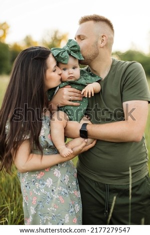 Young caucasian parents kissing pretty little daughter on cheeks from both sides, standing together in background of field. Happy family spending leisure time among green nature. Copy space.