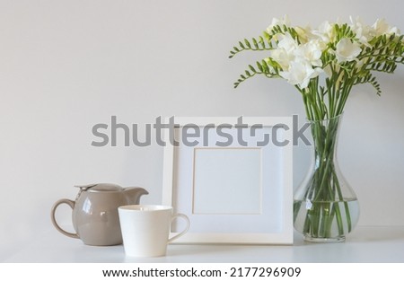 White freesia flowers in glass vase with blank square picture frame, tea cup and teapot against white background (selective focus)