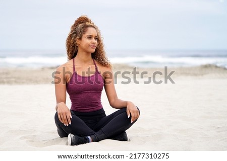 Beautiful, fit, young African American woman in work out clothing sitting cross legged at the beach in the sand, looking to the right                                Royalty-Free Stock Photo #2177310275