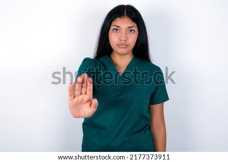 Doctor hispanic woman wearing surgeon uniform over white background shows stop sign prohibition symbol keeps palm forward to camera with strict expression