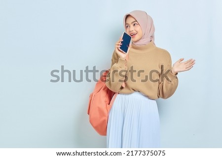 Cheerful young Asian Muslim woman student in brown sweater with backpack, singing his favorite song using mobile phone as microphone isolated on white background. back to school concept