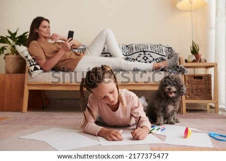 Girl lying on floor next to her dog and drawing pictures when mother reading in background