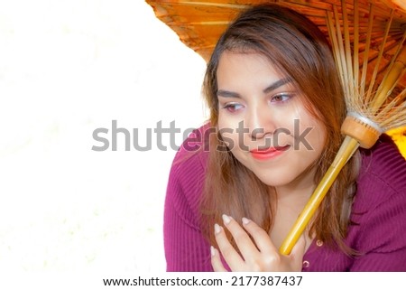 Portrait of young woman with slight smile looking down, holding yellow oriental umbrella, on white studio background, long straight brown hair, trendy eyebrows, natural makeup, casual violet blouse