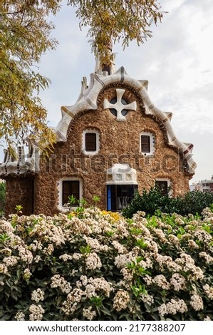 Vertical Photo of Porter's Lodge Pavillon in Park Güell. Catalan Landmark with Tree and Flower Bush on a Cloudy Day in Barcelona.
