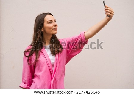 Portrait of a caucasian brunette smiling woman in casual pink shirt taking selfie, look at smartphone front camera, photographing, record video message post online. Isolated on a white background.