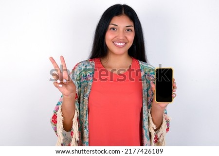 Young hispanic woman wearing colourful clothes over white background holding modern device showing v-sign