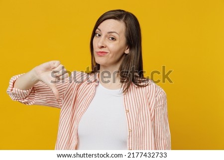 Young sad dissatisfied displeased unhappy woman she 30s in striped shirt white t-shirt showing thumb down dislike gesture isolated on plain yellow background studio portrait. People lifestyle concept Royalty-Free Stock Photo #2177432733