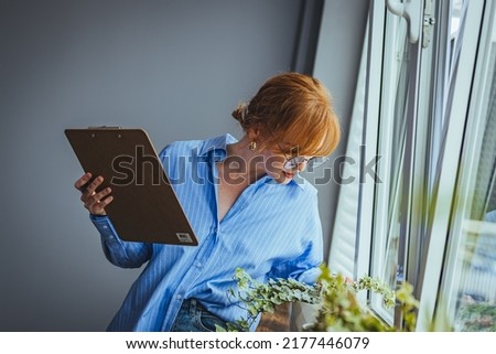 Busy beautiful female florist, owner of small business flower shop, checking stocks, inventory and data on clipboard against flowers and plants. Daily routine of running a small business
