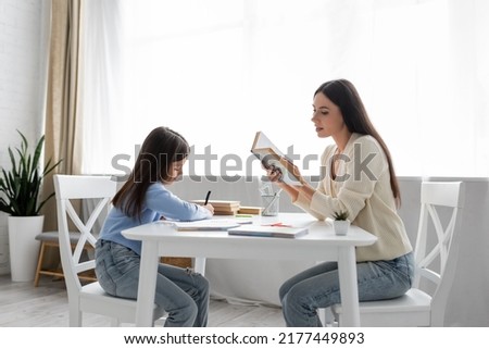 side view of babysitter reading aloud near girl writing dictation at home Royalty-Free Stock Photo #2177449893