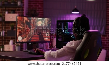 Over shoulder view of african american gamer girl playing online games using gaming pc setup spending relaxing time at home. Woman using modern technology for entertainment and streaming tournament. Royalty-Free Stock Photo #2177467491