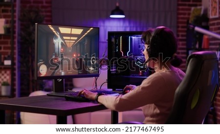 Gamer girl using pc setup playing multiplayer first person shooter talking on headset while explaining gameplay to subscribers. Woman streaming online action game while sitting in gaming chair.