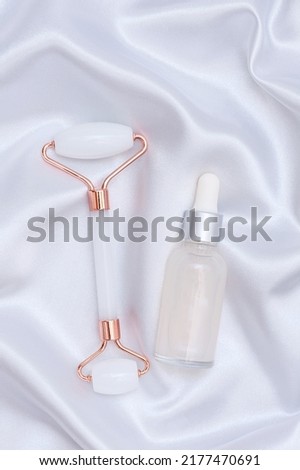 A glass bottle with a pipette-dropper with serum or essential oil and a massage roller for the face on a white silk fabric in waves. vertical photo