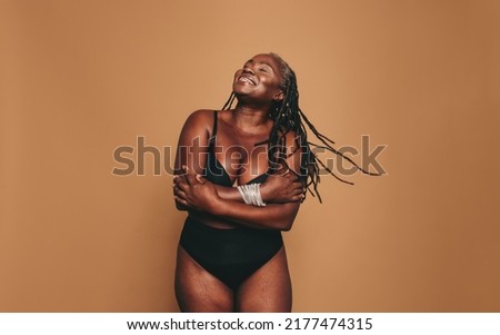 Loving my natural body. Happy woman with dreadlocks embracing her natural and ageing body. Mature black woman smiling cheerfully while standing against a studio background in black underwear. Royalty-Free Stock Photo #2177474315
