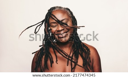 Cheerful woman smiling and whipping her dreadlocks while standing against a studio background. Mature black woman wearing a bath towel and make-up. Happy middle-aged woman embracing her ageing body. Royalty-Free Stock Photo #2177474345