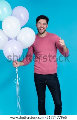 Positive Smiling Funny Caucasian Guy Handsome Brunet Man With Bunch of Colorful Air Balloons in Pink Jumper Showing Thumbs Up Against Blue. Vertical Image