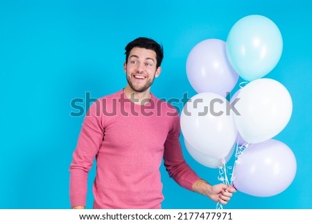 Smiling Happy Caucasian Guy Handsome Brunet Man With Bunch of Colorful Air Balloons in Pink Jumper Standing On Blue Background. Horizontal image