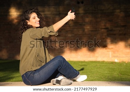 Young and beautiful latin woman with curly brown hair and dressed in casual clothes sitting on a grey wall taking pictures of herself with her mobile phone while smiling. Concept of technology.