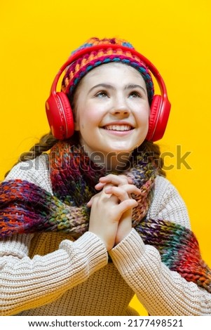 One Winsome Relaxed and Smiling Caucasian Blond Girl In Warm Knitted Hat and Scarf While Listening to Music in Wireless Headphones Over Yellow. Vertical Shot