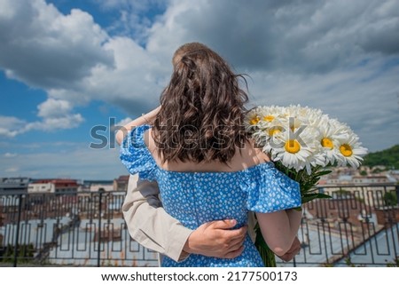 Man hugs and gently supports girl in a blue dress with daisies from behind. The background is a view of the city. Without faces