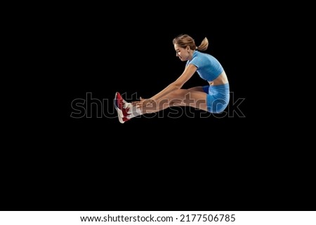 Side view. Young sportive girl, long jumper in sports blue uniform performs triple jump isolated on black background. Concept of sport, action, motion, speed, healthy lifestyle. Copy space for ad Royalty-Free Stock Photo #2177506785