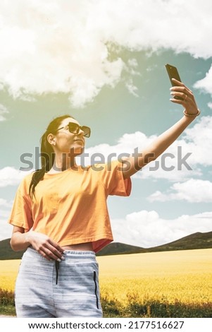 girl on the road, in a yellow T-shirt, taking a photo in a flowering field, against the backdrop of mountains