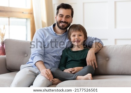 Portrait of smiling young Caucasian single father sit on sofa in new house with little son. Happy loving dad and small boy child renters tenants relax on couch at home, enjoy family weekend together.