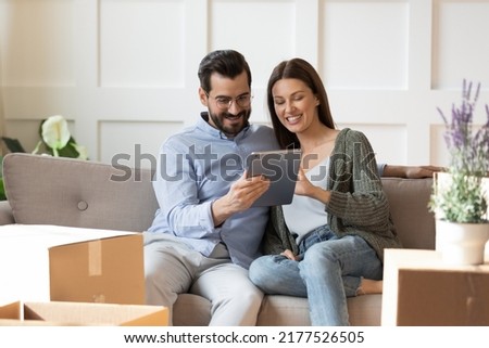 Smiling young Caucasian couple relax on sofa in living room on moving day watch video on tablet. Happy man and woman renters tenants rest on couch relocate to new home, use pad browsing internet. Royalty-Free Stock Photo #2177526505